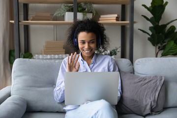 Happy millennial African American woman in headphones wave at camera talk on video call on laptop at home. Smiling young biracial female in earphones have webcam communication on computer.