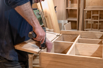 hands of a master carpenter install the hinges on the cabinet with a hand-held power tool screwdriver. Production of handmade wooden furniture. Selective focus close-up