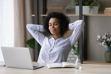 Calm millennial African American businesswoman relax at desk at home office breathe fresh air. Young mixed race woman sit at table work on computer take nap or sleep, relieve negative emotions.