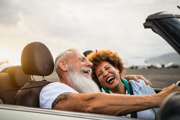 Happy senior couple having fun driving on new convertible car - Mature people enjoying time together during road trip tour vacation - Elderly lifestyle and travel culture transportation concept - Powered by Adobe