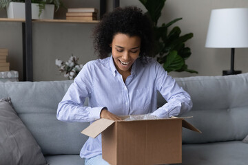 Overjoyed millennial African American woman open unpack box with internet order shopping online form home. Happy young biracial female excited with good quality delivery service. Shipping concept.