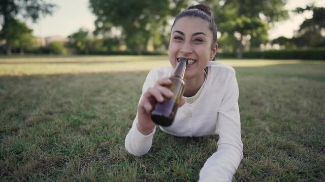 Pov shot of cute young gen z brunette girl talking to followers in a clip for social network lying on a city park drinking a beer for toasting online. Person in vacation outdoor video conferencing