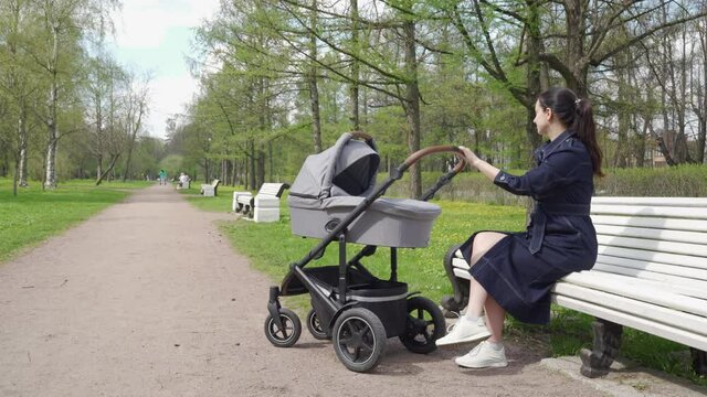 Mother with baby stroller on a walk in spring park, woman in denim dress with baby carriage sits on a bench in the park. High quality 4k footage