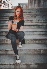 young woman or teenage girl eating asian fast food from takeaway box sitting on the steps on city street. Thai noodles in paper box takeaway street food