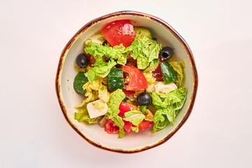 Greek salad of lettuce leaves, tomatoes, cucumbers, olives, cheese and butter in a white round plate on a white background.