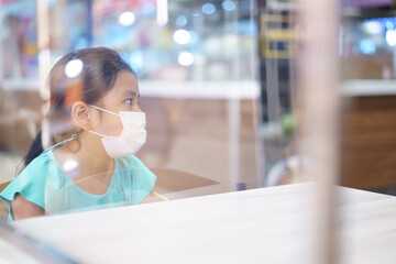 Asian child or kid girl wearing white face mask sitting behind protection glass partition and absent-minded on table for protect coronavirus covid-19 to social distancing and unhappy at food court