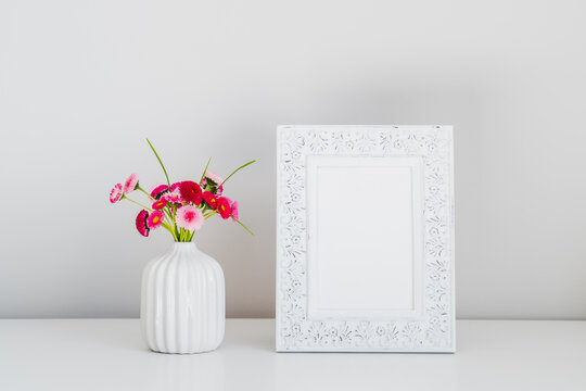 Photo frame mockup and boquet of pink spring flowers in vase on table on white background. Minimal style. Floral home decor.