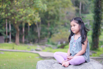 Asian child or kid girl open eyes mindfulness meditating with breath and sit on stone or rock for peace and relax or yoga meditation on nature green garden and tree at temple park or church outdoor