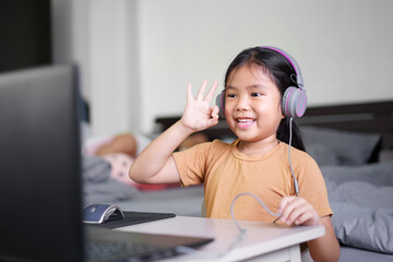 Asian child student or kid girl smile enjoy and make hand ok on computer notebook and wearing headphone to listening communication on study online or people learn from home on laptop by back to school