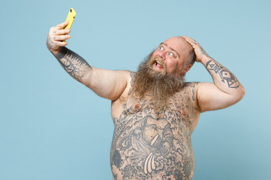 Young funny fat pudge obese chubby overweight blue-eyed bearded man 30s has big belly with naked tattooed torso doing selfie shot on mobile cell phone hold head isolated on pastel blue background