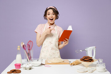 Young amazed fun housewife housekeeper chef baker woman in pink apron work at table kitchenware point on recipe cook book look aside isolated on pastel violet background Process cooking food concept. - Powered by Adobe