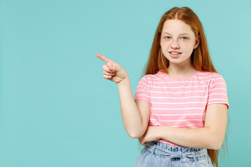 Little fun redhead kid girl 12-13 years old in pink striped t-shirt point index finger aside on workspace area mock up isolated on pastel blue background studio. Children lifestyle childhood concept.