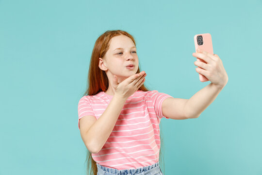 Little redhead kid girl 12-13 years old wearing pink striped t-shirt do selfie shot on mobile phone post photo social network blow kiss isolated on pastel blue background Lifestyle childhood concept.