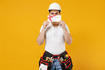 Fototapeta na wymiar Young smiling employee handyman man in protective helmet hardhat cover eye with gift voucher flyer mock up isolated on yellow background Instruments for renovation apartment room Repair home concept.