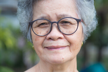 Portrait of an elderly woman with short gray hair, wearing glasses, smiling, and looking at the camera while standing in a garden. Aged people and relaxation concept