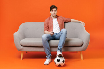 Young happy fun man football fan in shirt support favorite team with soccer ball sit on sofa at...