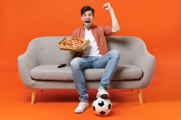 Young man football fan in shirt support favorite team with soccer ball sit on sofa at home watch tv live stream eat pizza do winner gesture isolated on orange background People sport leisure concept - obrazy, fototapety, plakaty