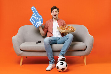 Young man football fan in shirt support team with soccer ball sit on sofa at home watch tv live stream hold pizza fan foam glove finger isolated on orange background People sport lifestyle concept