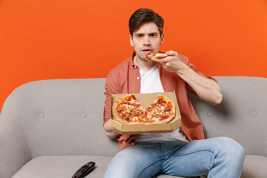 Young serious caucasian man 20s wearing shirt sit on gray sofa at home watching tv live stream movie film eat pizza rest isolated on orange background studio portrait People leisure lifestyle concept.