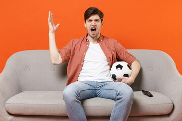 Fototapeta Young indignant angry man football fan wearing shirt support team with soccer ball sitting on sofa home watching tv live stream spread hand isolated on orange background studio. People sport concept obraz