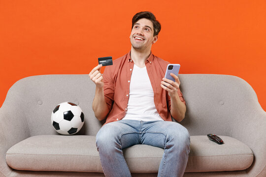 Young Dreamful Happy Man Football Fan Wearing Shirt Support Team With Soccer Ball Sit On Sofa Home Watch Tv Hold Mobile Cell Phone Credit Bank Card Isolated On Orange Background. People Sport Concept.