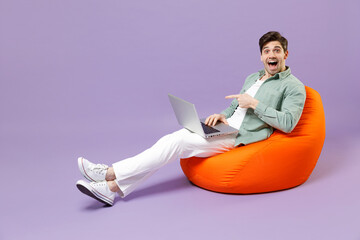 Full length fun man 20s in casual mint shirt white t-shirt sitting in orange bean bag chair point finger on laptop pc computer chat isolated on purple color background studio People lifestyle concept.