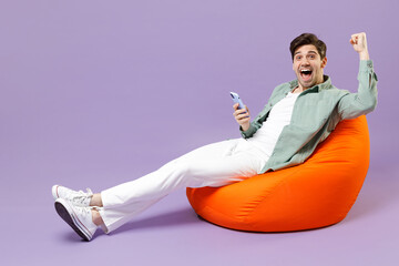 Full length man in casual mint shirt white t-shirt sitting in orange bean bag chair use mobile cell...
