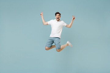 Obraz premium Full length smiling young man 20s wear white casual basic t-shirt showing victory v-sign gesture jump high looking camera isolated on pastel blue background studio portrait. People lifestyle concept.