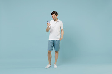 Fototapeta na wymiar Full length young smiling man 20s wearing white casual basic t-shirt hold mobile cell phone chat online browsing internet isolated on pastel blue background studio portrait People lifestyle concept.