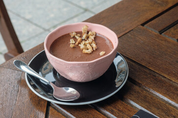 A pink bowl with chocolate mousse, pieces of walnut and a teaspoon on the dark brown table at cafe
