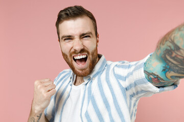 Close up young caucasian unshaven man 20s in blue striped shirt doing selfie shot on mobile cell phone clench fist do winner gesture isolated on pastel pink background studio People lifestyle concept