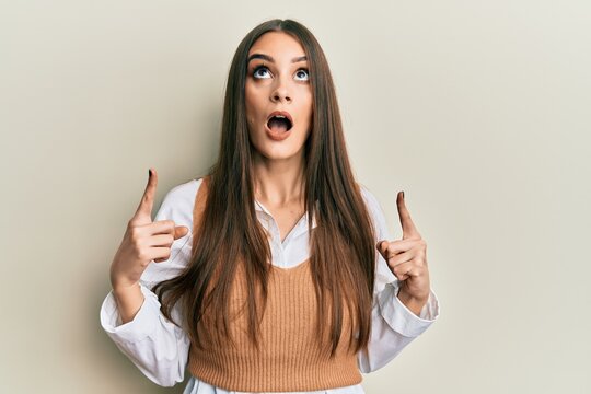 Beautiful brunette young woman wearing casual clothes amazed and surprised looking up and pointing with fingers and raised arms.