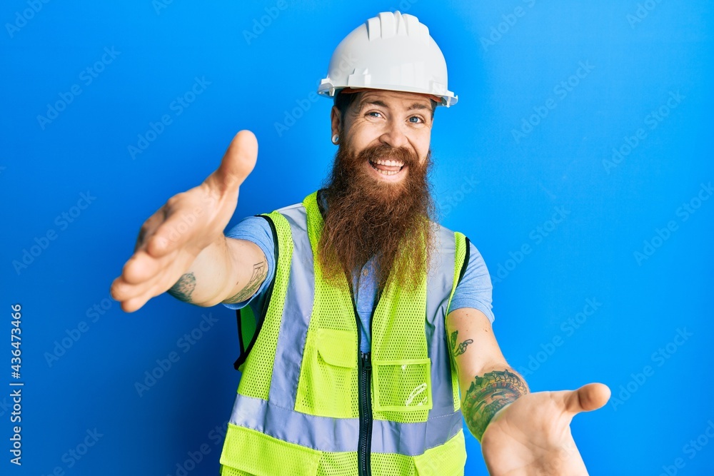 Poster redhead man with long beard wearing safety helmet and reflective jacket looking at the camera smilin