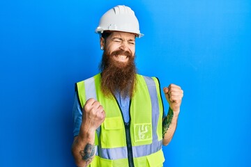 Redhead man with long beard wearing safety helmet and reflective jacket excited for success with...