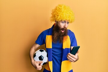 Redhead man with long beard football hooligan cheering game holding smartphone afraid and shocked with surprise and amazed expression, fear and excited face.