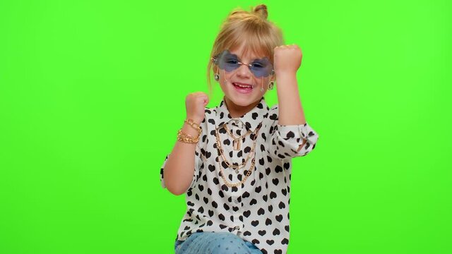 Little blonde teen kid child girl in shirt celebrate success win scream rejoices doing winner hands gesture say Yes, dancing isolated on chroma key green background. Young children lifestyle emotions