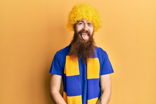 Redhead man with long beard football hooligan cheering game wearing funny wig winking looking at the camera with sexy expression, cheerful and happy face.
