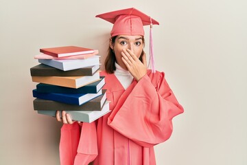 Young caucasian woman wearing graduation cap and ceremony robe covering mouth with hand, shocked and afraid for mistake. surprised expression