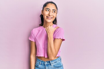 Hispanic teenager girl with dental braces wearing casual clothes pointing thumb up to the side smiling happy with open mouth