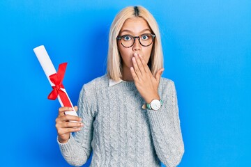 Beautiful blonde woman holding graduate degree diploma covering mouth with hand, shocked and afraid...