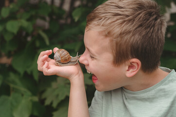 A happy laughing kid holds a big garden snail and touches it with his finger in the park