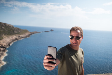 Happy young adult smiling and wearing sunglasses holding a smartphone and taking a selfie with the ocean in the background. 