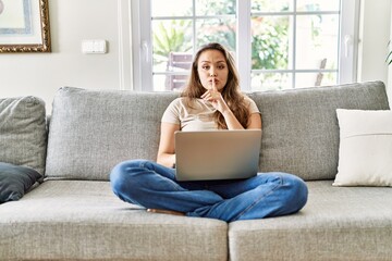 Beautiful young brunette woman sitting on the sofa using computer laptop at home asking to be quiet...