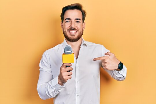 Handsome caucasian man with beard holding reporter microphone pointing finger to one self smiling happy and proud