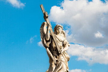 A stone statue of an angel with wings holding a holy cross in Rome, Italy with a blue sky...