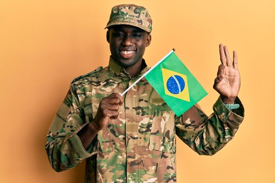 Young african american man wearing army uniform holding brazil flag doing ok sign with fingers, smiling friendly gesturing excellent symbol