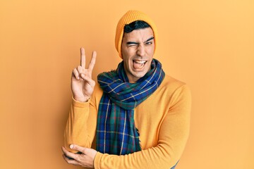 Handsome hispanic man wearing wool hat and winter scarf smiling with happy face winking at the...