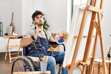 Young hispanic man sitting on wheelchair painting at art studio pointing up looking sad and upset,...