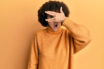 Young hispanic girl wearing wool winter sweater peeking in shock covering face and eyes with hand,...