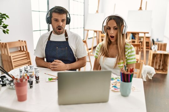Young couple of wife and husband at art studio looking at video on laptop scared and amazed with open mouth for surprise, disbelief face
